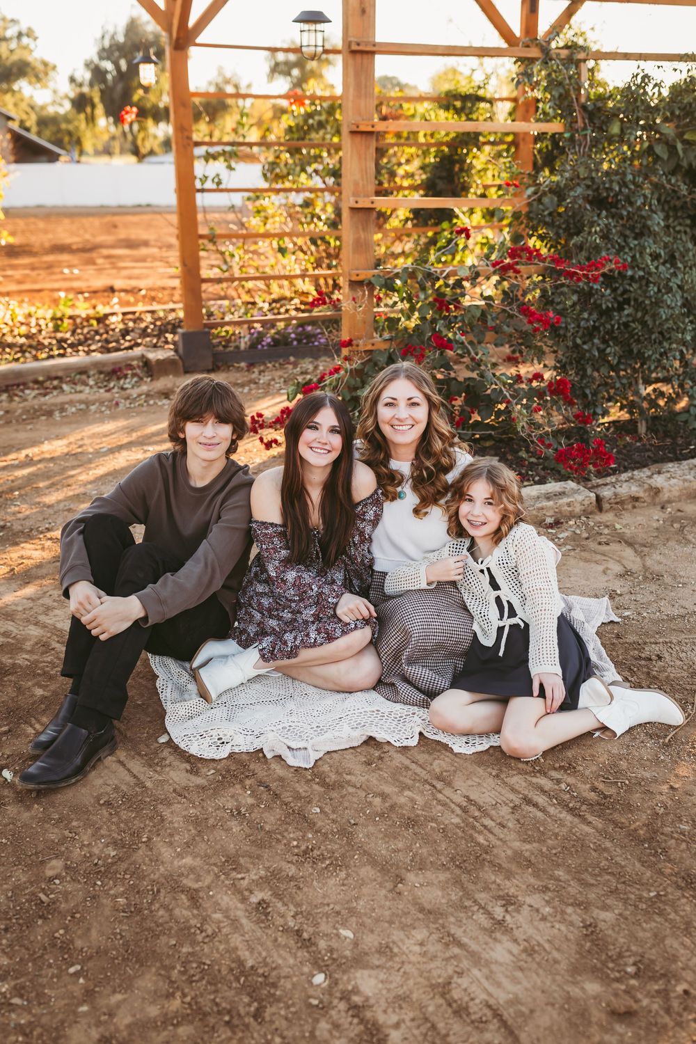 A mom and her kids together on a blanket at sunset with wooden pergola and flowering plants in background.