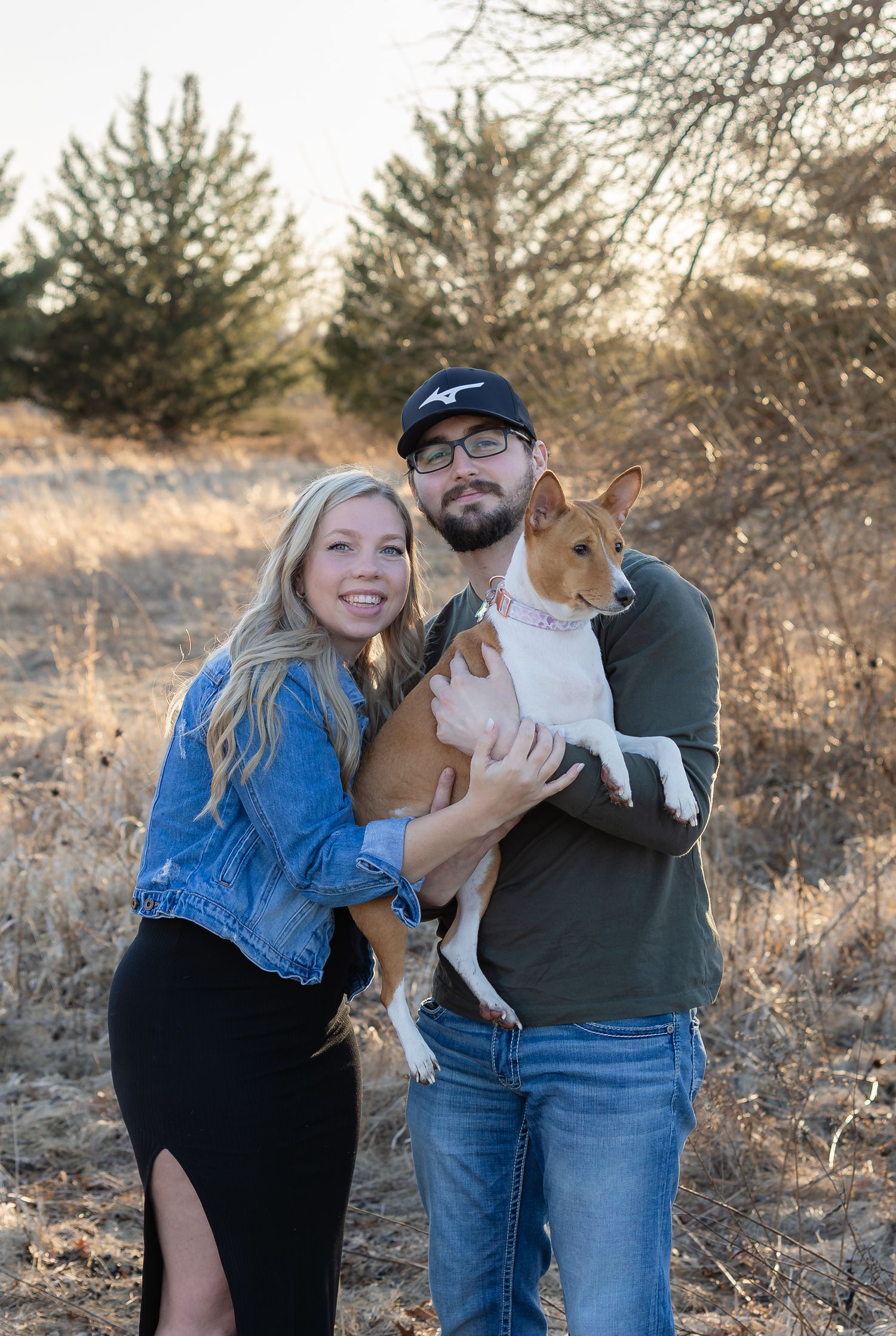 A couple holds their brown and white dog while posing together outdoors in a natural setting during golden hour.