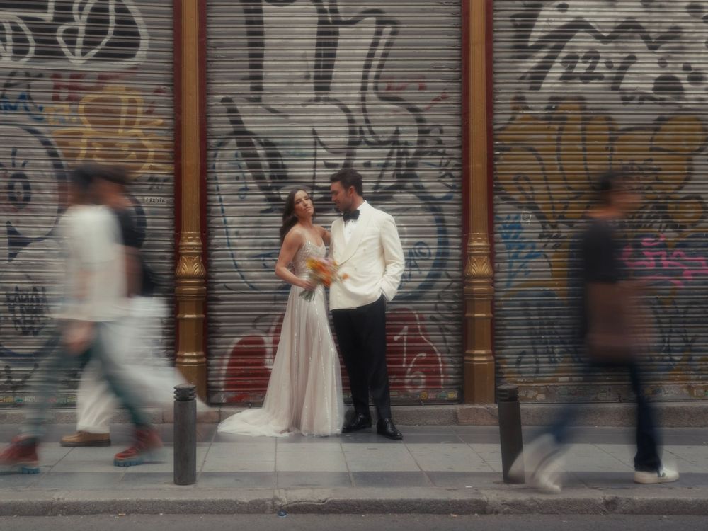 People walk past a wall covered in graffiti and closed storefront shutters on an urban street.
