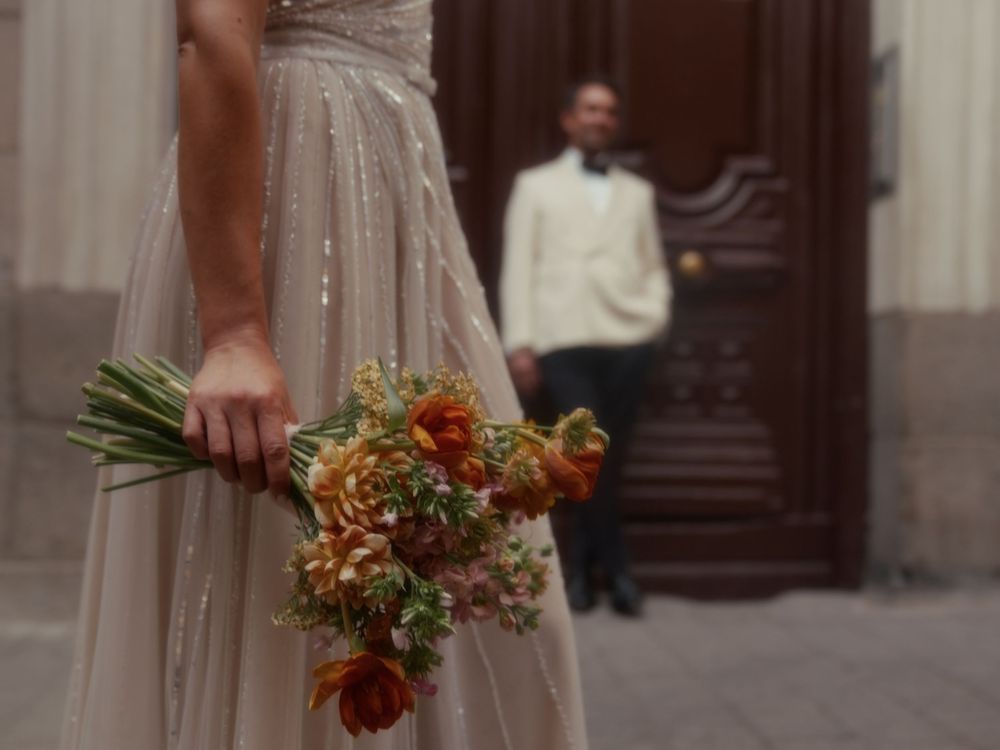 Wedding party members hold autumn-themed bouquets with orange and burgundy flowers against a stone building backdrop.