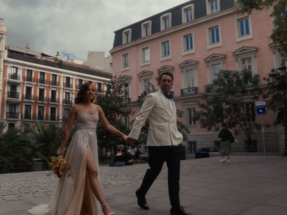 A couple dances together in a romantic moment on a European town square with pink buildings in the background.