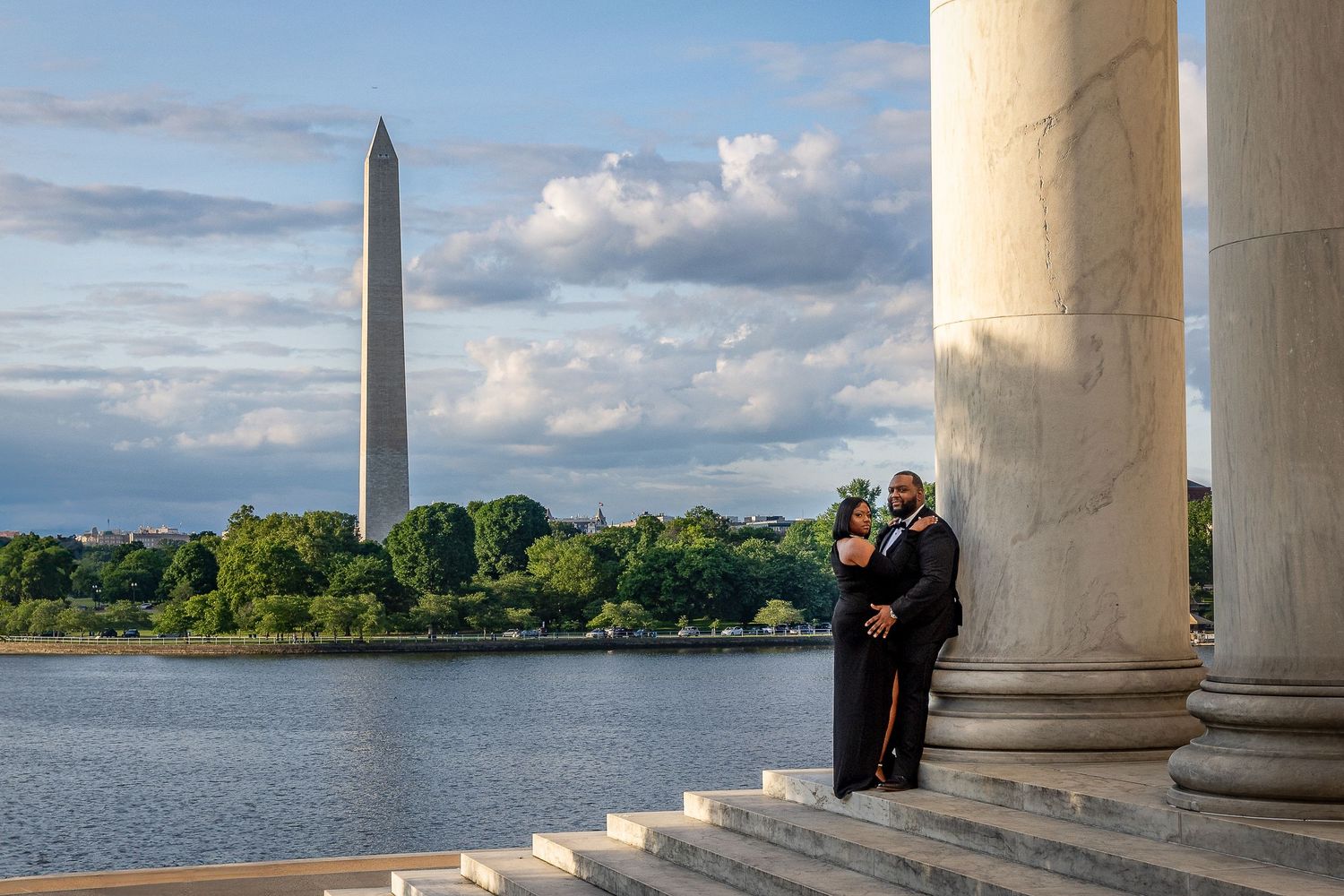 A couple stands between marble columns at the Jefferson Memorial with the Washington Monument visible across the Tidal Basin.