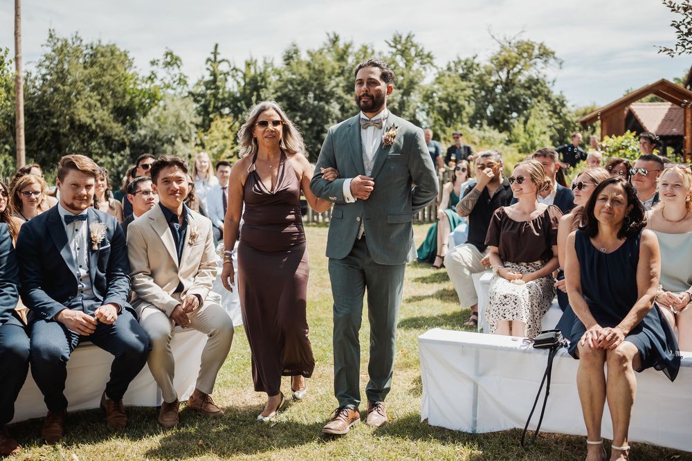 Outdoor wedding celebration with guests gathered on a sunny lawn near a rustic building.