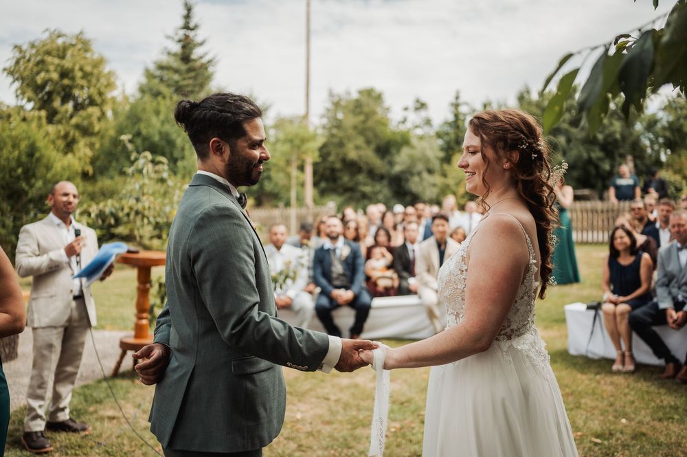 Romantic outdoor wedding ceremony with guests seated on wooden benches surrounded by lush greenery and trees.