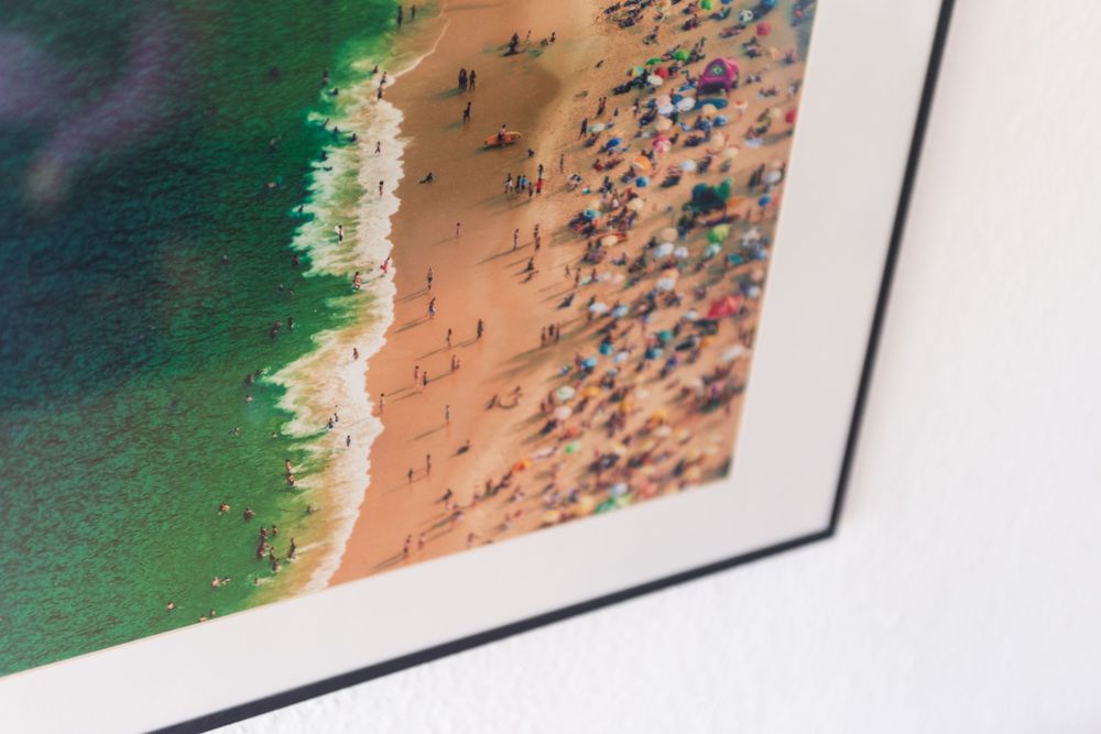 Quadro com moldura preta, exibindo fotografia aérea e colorida de uma praia cheia de guarda-sóis e pessoas. Solicite orçamento para quadros.