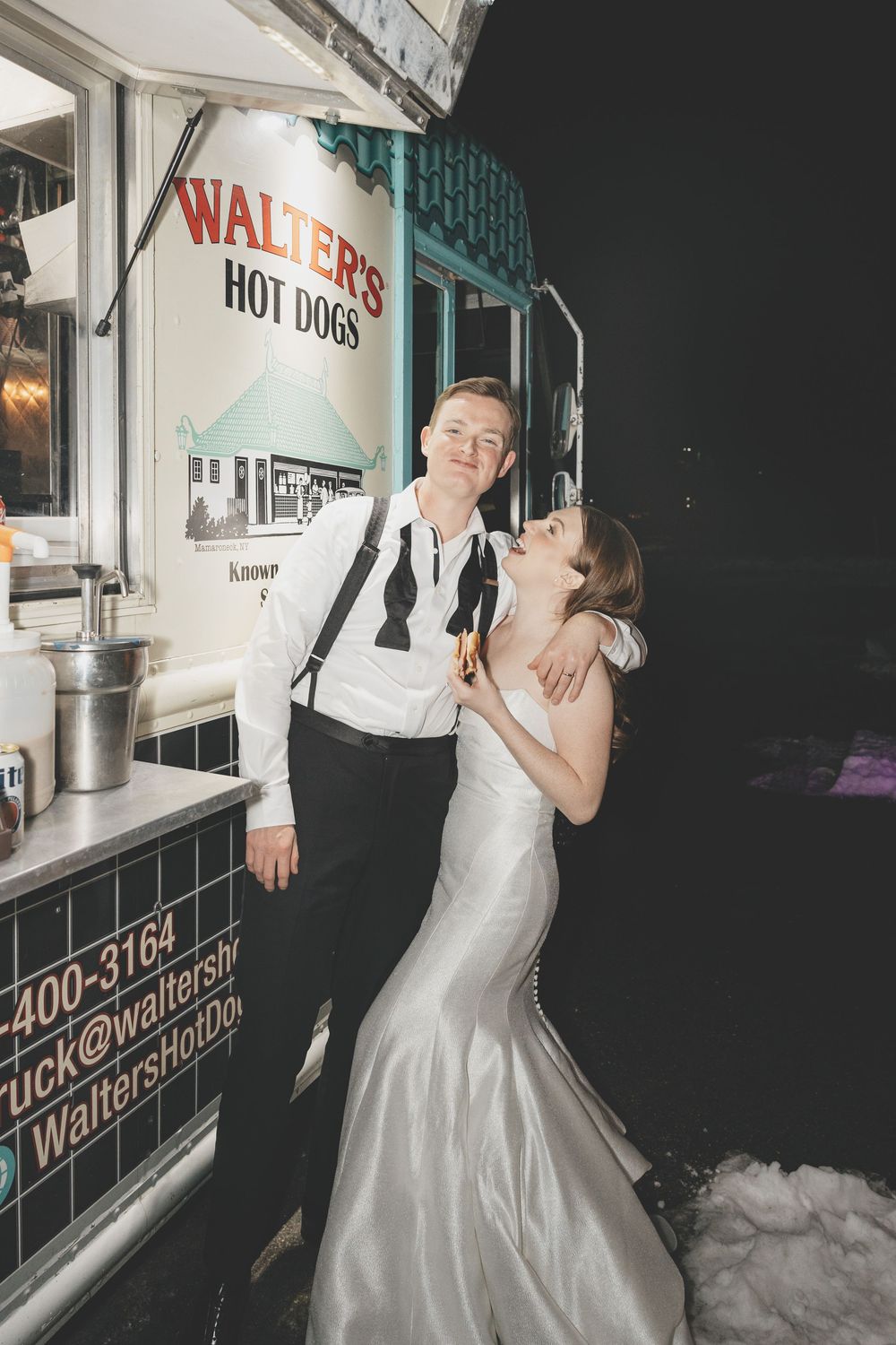 A couple shares an intimate moment at night near an illuminated food truck with 'Hot Dogs' signage.