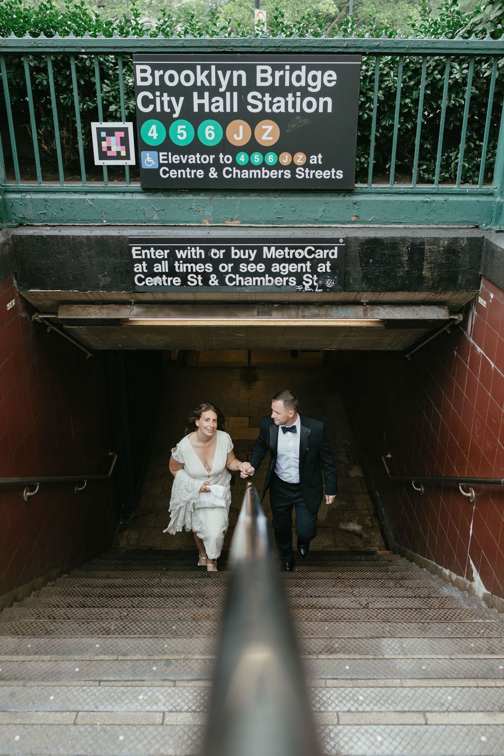 A couple in formal wedding attire descends the stairs at Brooklyn Bridge City Hall subway station entrance.