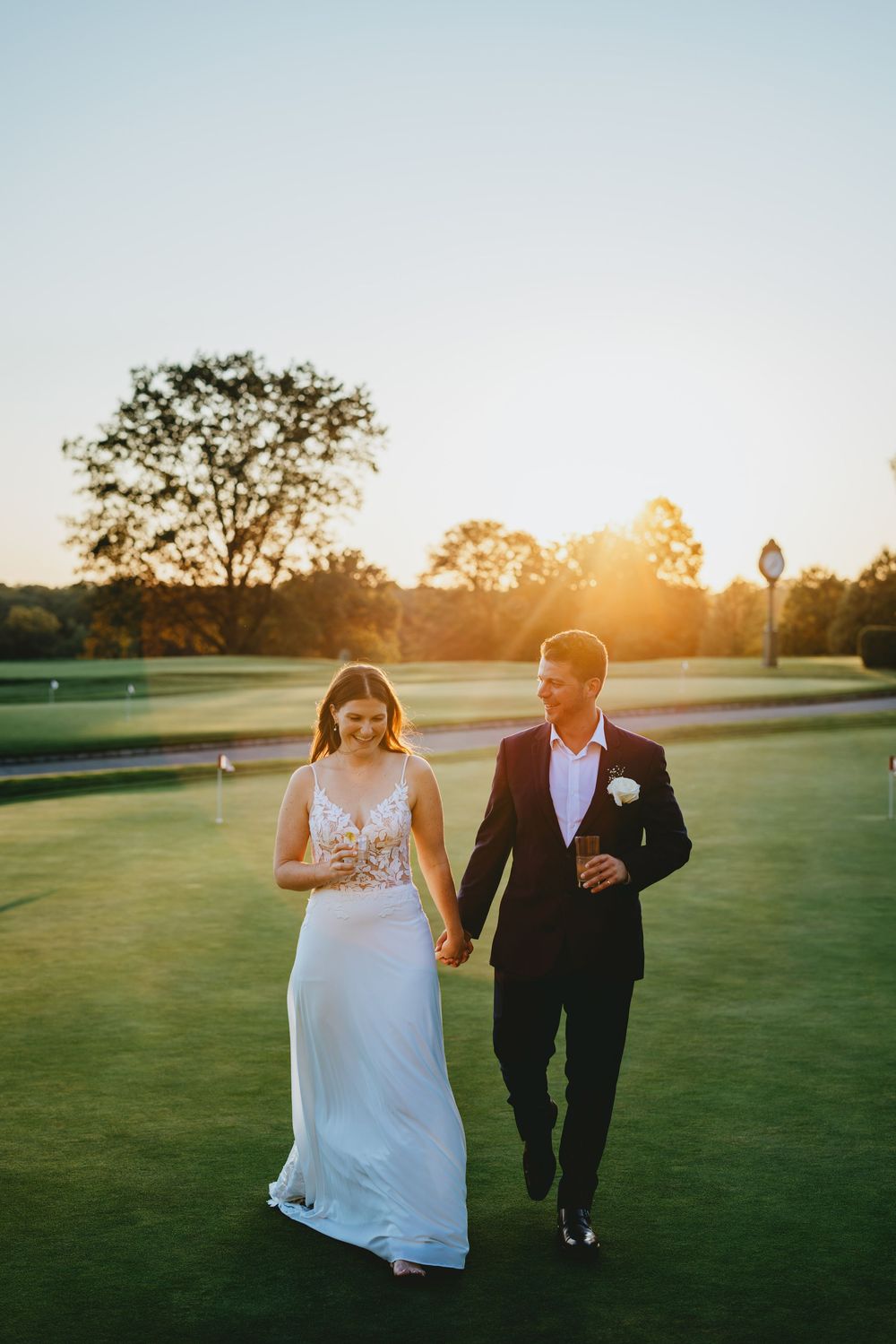 A couple walks hand in hand across a golf course at sunset with golden light illuminating their formal wedding attire.