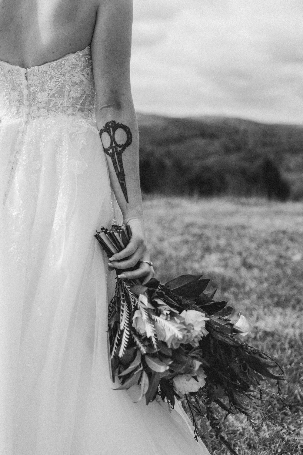 Black and white photo of a bridal bouquet with feathers and flowers held against a grassy outdoor landscape.
