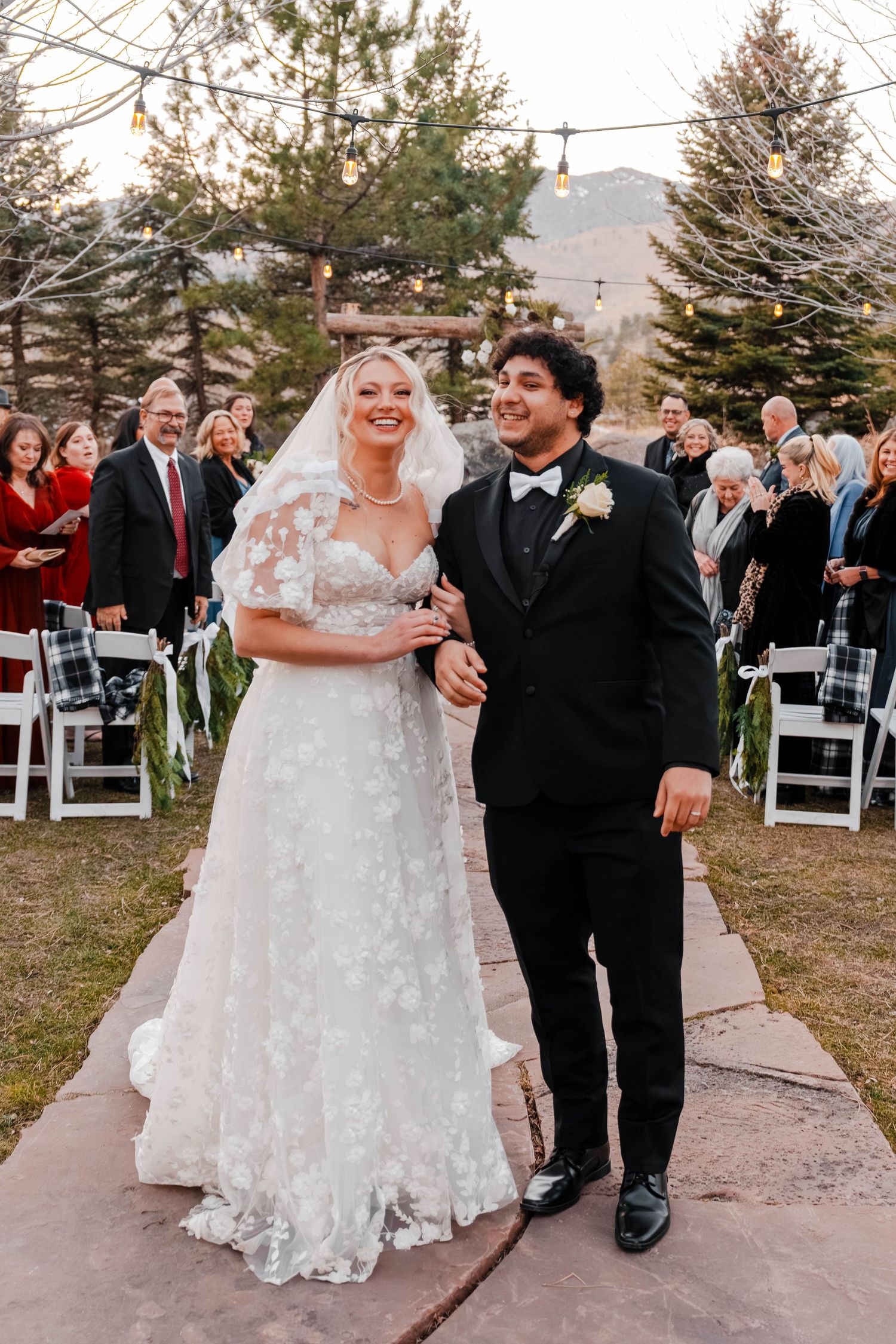 Bride and groom walking down outdoor wedding aisle surrounded by guests at dusk.