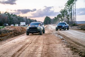 A Black Dodge Ram TRX and Blue Ford Raptor  race along a dusty dirt road during sunset in Charleston, SC.