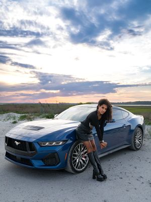 A model posing in front of a blue Ford Mustang parked on Folly Beach, SC at sunset with dramatic cloudy skies overhead.