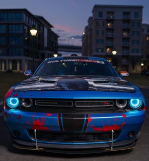 A modified Dodge Challenger with glowing blue headlights and red-blue racing stripes parked at dusk between city buildings,Charleston, SC.