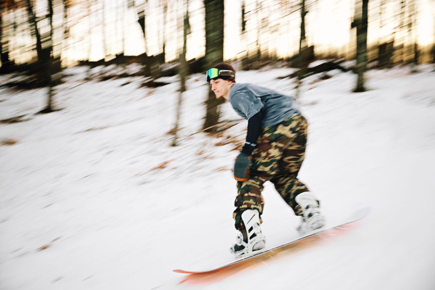 Snowboarder in camo pants and green goggles speeding down a snowy slope through a wooded trail.