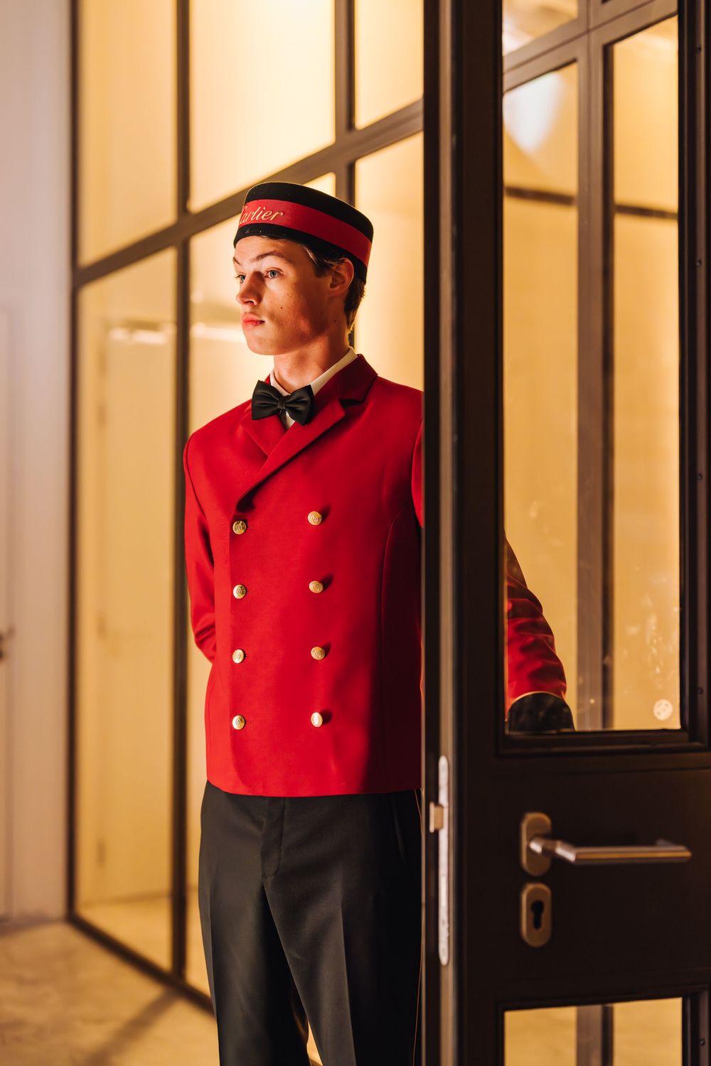Elevator operator in traditional red uniform stands in vintage elevator with glass panels and brass details.