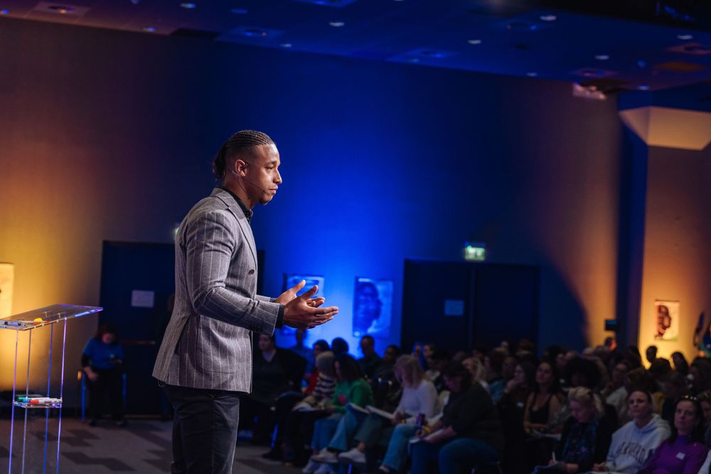 A speaker delivers a presentation on stage with dramatic blue lighting at an event venue.