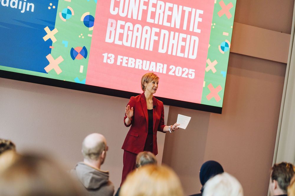 A speaker presents at a Dutch conference with projection screen displaying event details.