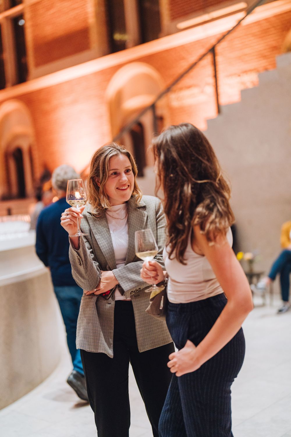 Two women enjoying conversation and wine at an elegant indoor event with warm lighting and architectural details.