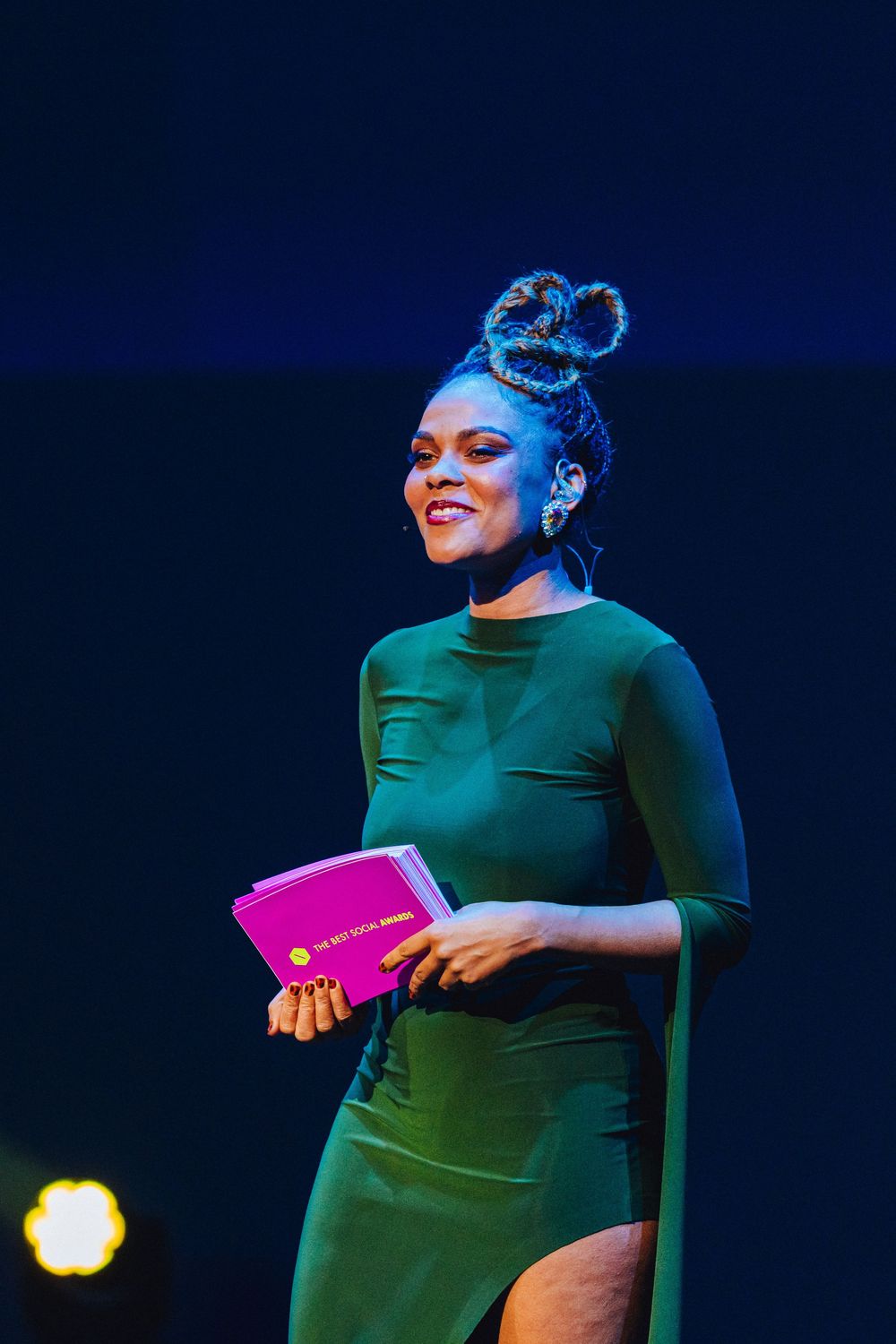 A performer in an emerald green dress holds a pink book while standing on stage under blue lighting.