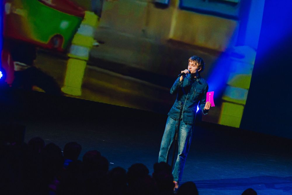 A performer in a blue denim jacket stands on stage with dramatic blue and yellow lighting effects during a concert.