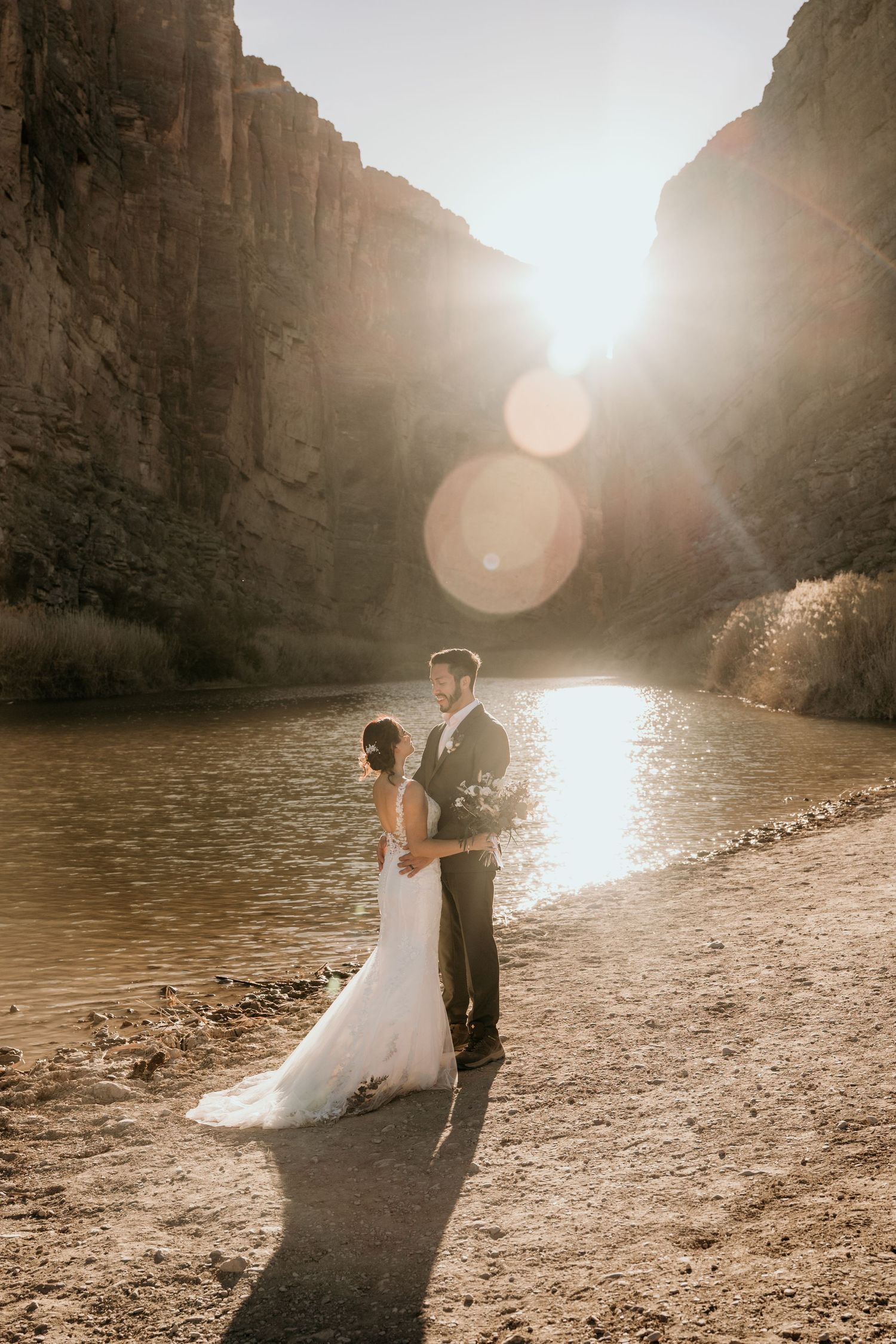 Wedding couple dances on sandy beach as sun sets behind canyon walls.