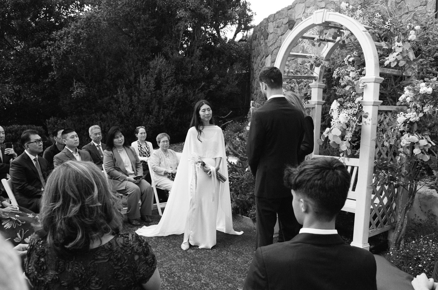 Black and white outdoor wedding ceremony with bride in white gown, groom, and seated guests beside a floral arch.