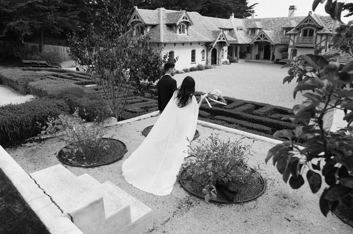 Bride and groom walk through elegant garden grounds of a historic manor house in black and white wedding photo.