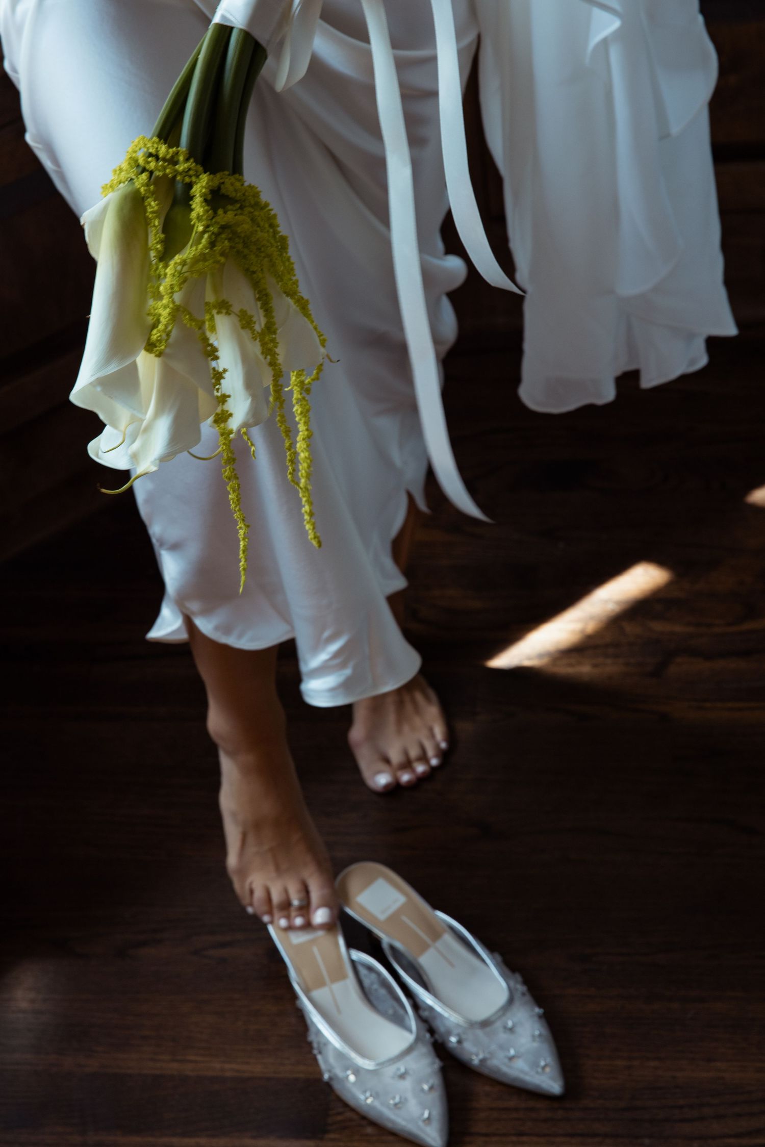 White flowy dress and bare feet next to delicate beaded bridal shoes on dark wooden floor with yellow flowers.