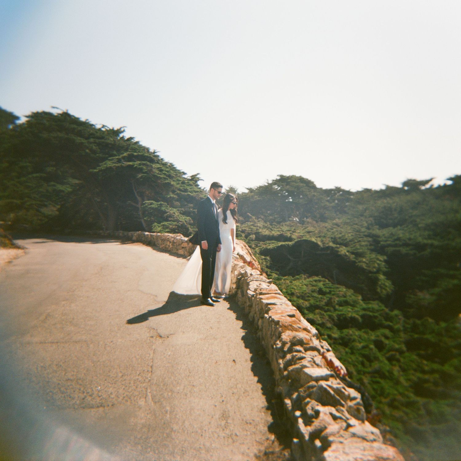 Bride and groom standing on a stone bridge surrounded by lush green cypress trees on their wedding day.