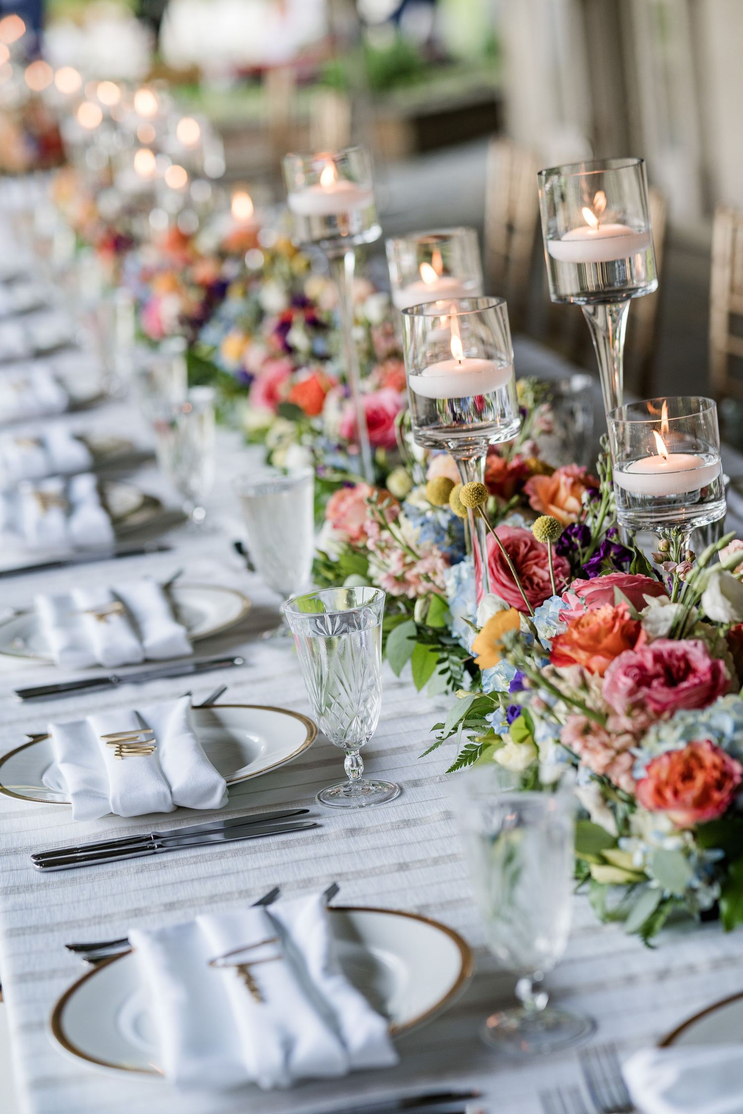 Elegant wedding table setting with floating candles, colorful floral centerpieces, and white napkins on a long banquet table.