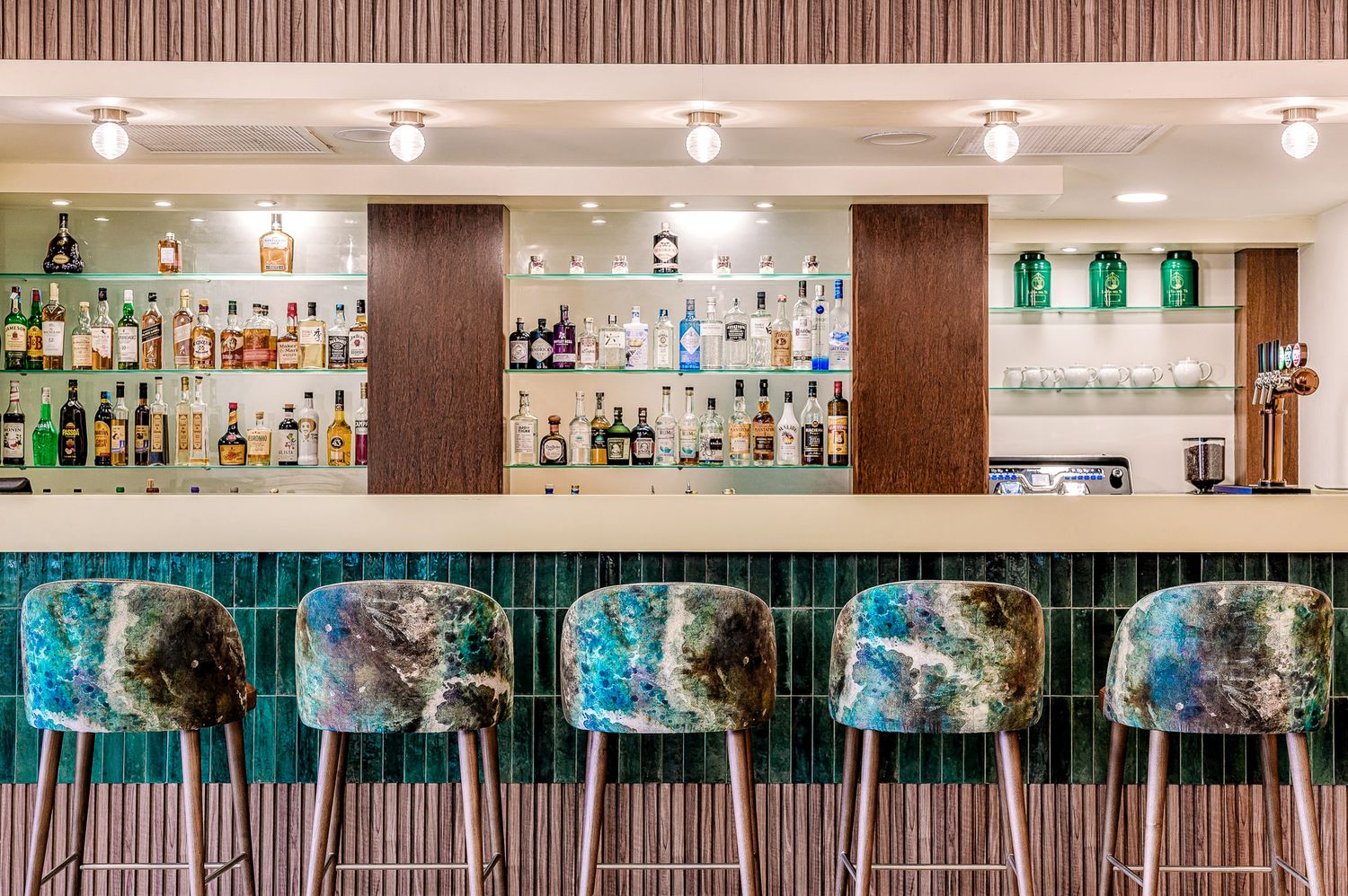 A modern bar interior with illuminated shelves displaying bottles of liquor and glassware against a wood-paneled wall.