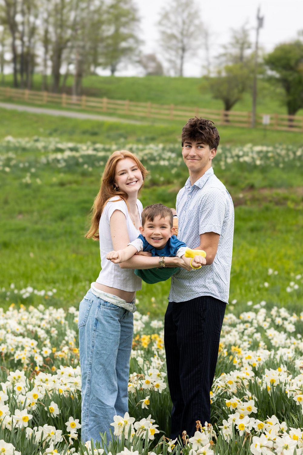 A family poses together in a spring field filled with blooming daffodils and white flowers on a green hillside.