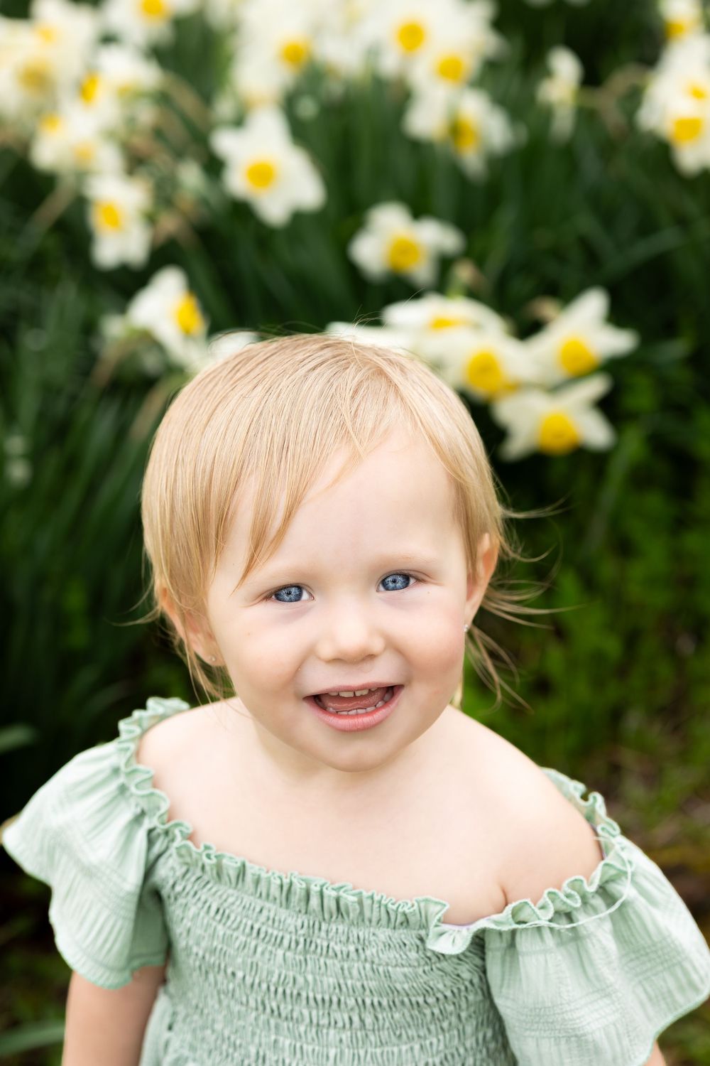 Child in mint green smocked dress smiles joyfully among blooming spring daffodils.