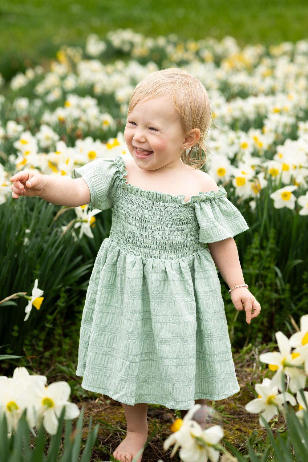 A small child in a mint green dress plays among blooming white daffodils in a spring garden.