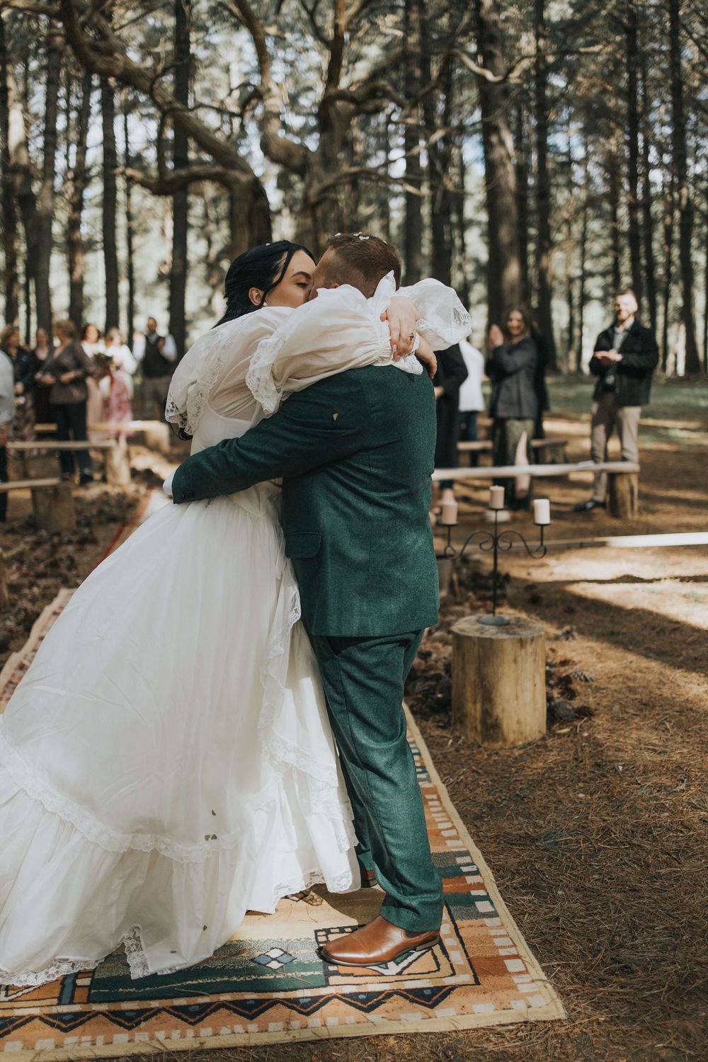 Bride and groom sharing their first kiss at an outdoor forest wedding ceremony with guests watching.