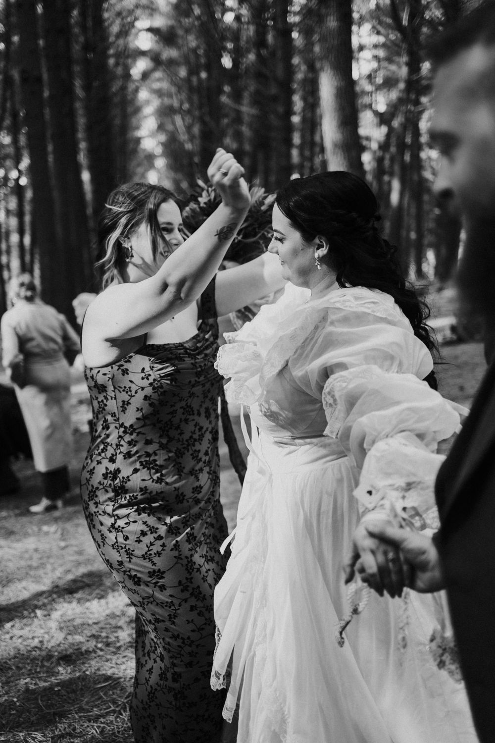Black and white photo of bride and bridesmaid dancing joyfully together in a pine forest wedding reception.