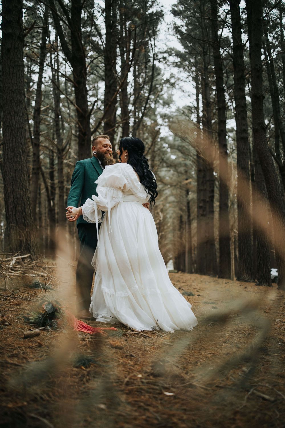 Bride in flowing white tiered gown dances with groom in green suit on a pine forest floor.