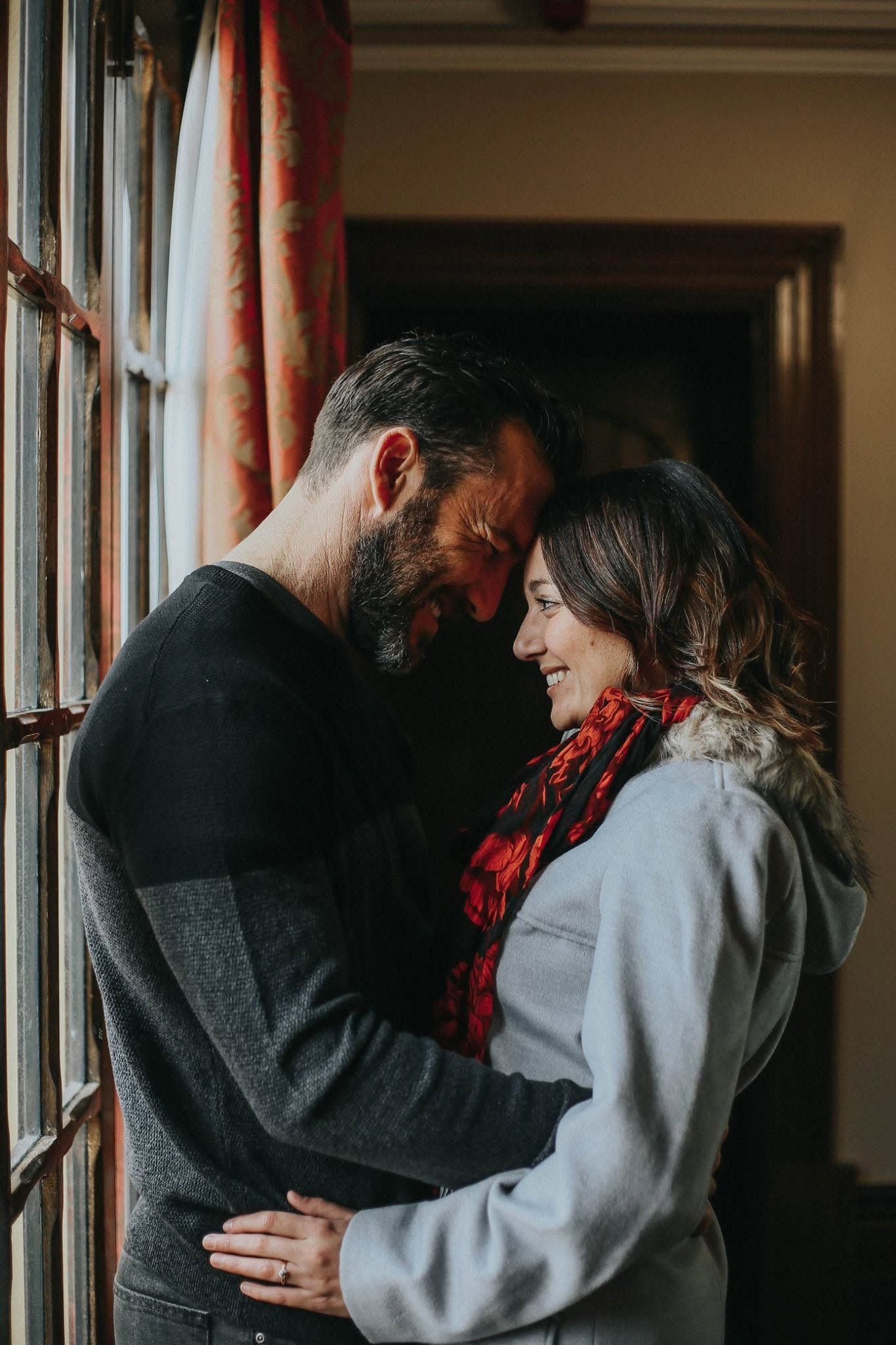 A couple shares a tender forehead-to-forehead embrace near a window, woman in red scarf smiling warmly at bearded man.