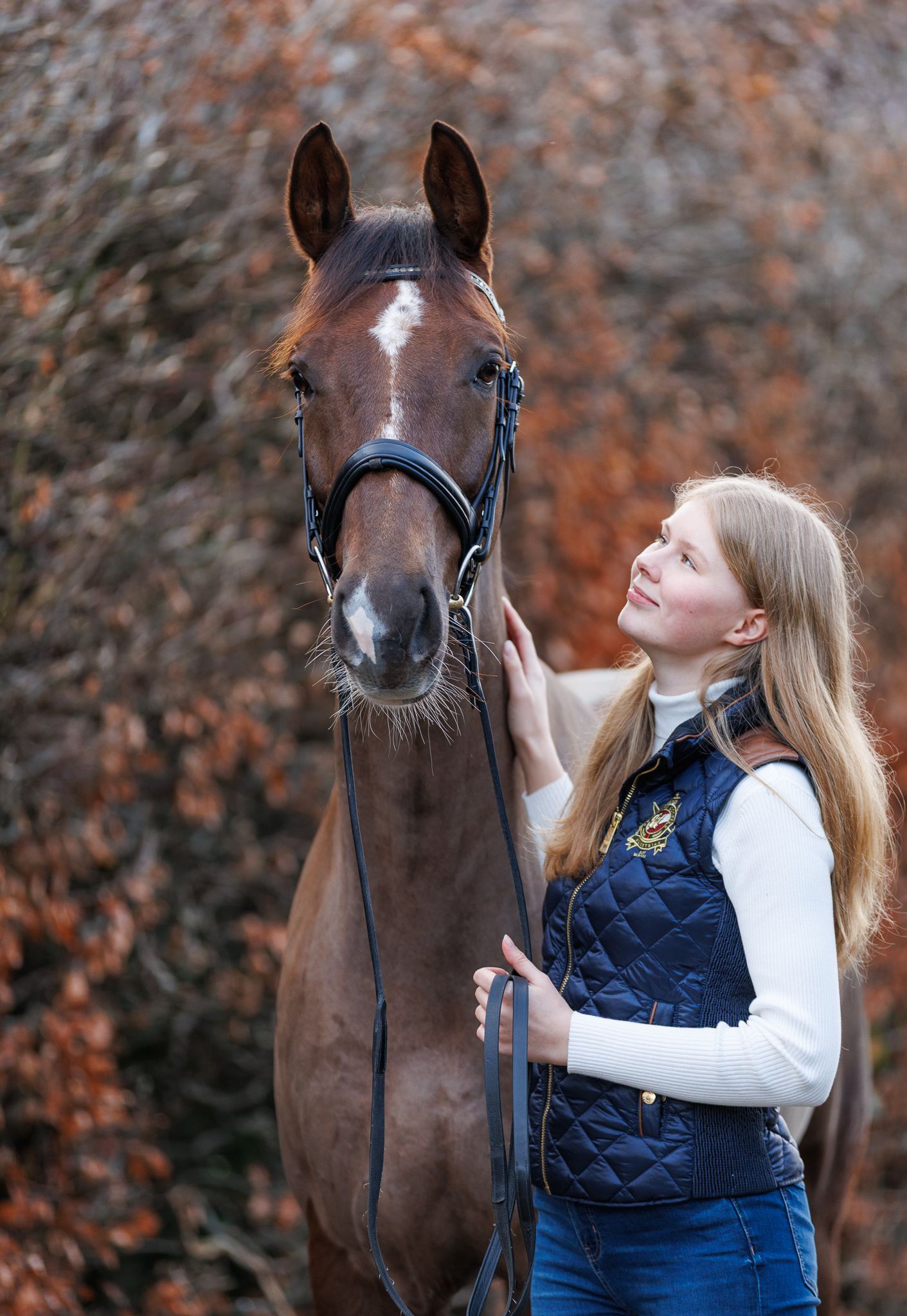 A beautiful bay horse wearing a black bridle stands next to someone in a white lace dress in an outdoor setting.