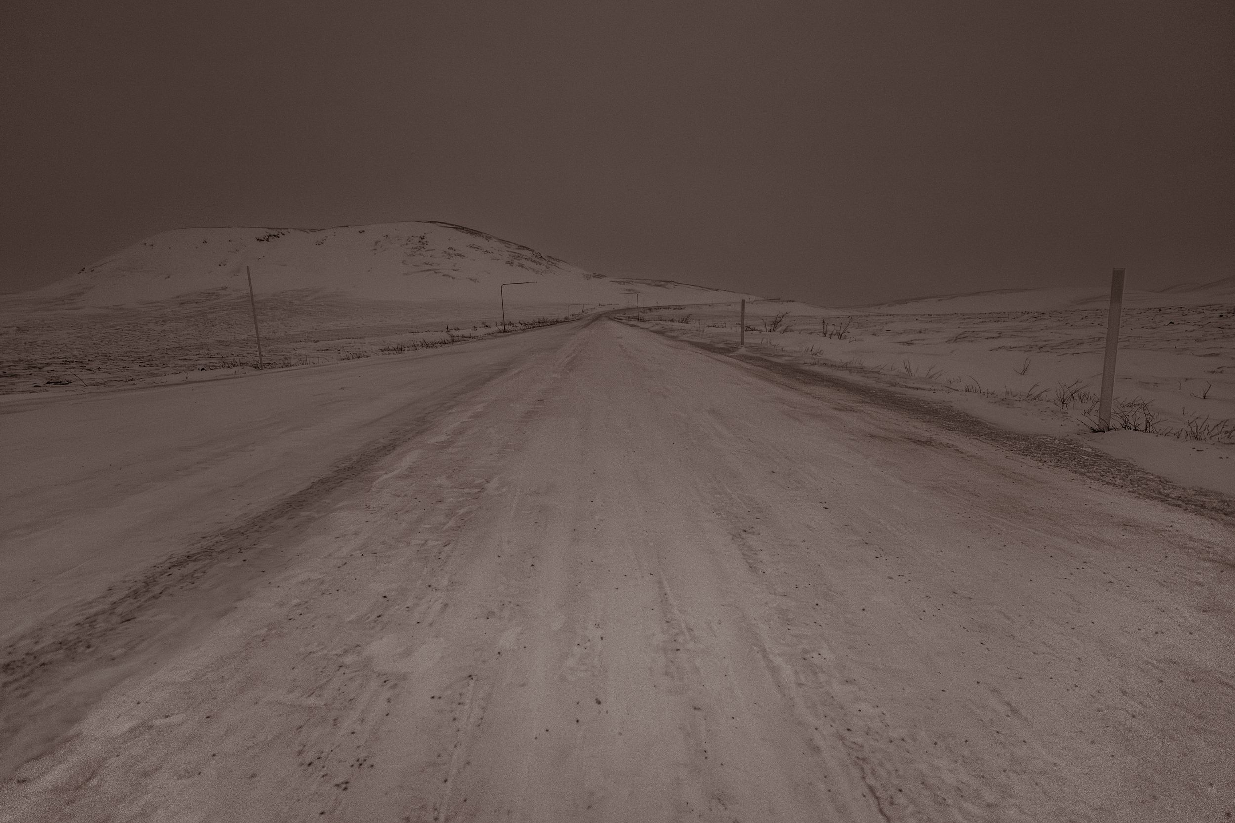 A snowy rural road stretches into the distance with power lines and fence posts visible along both sides.