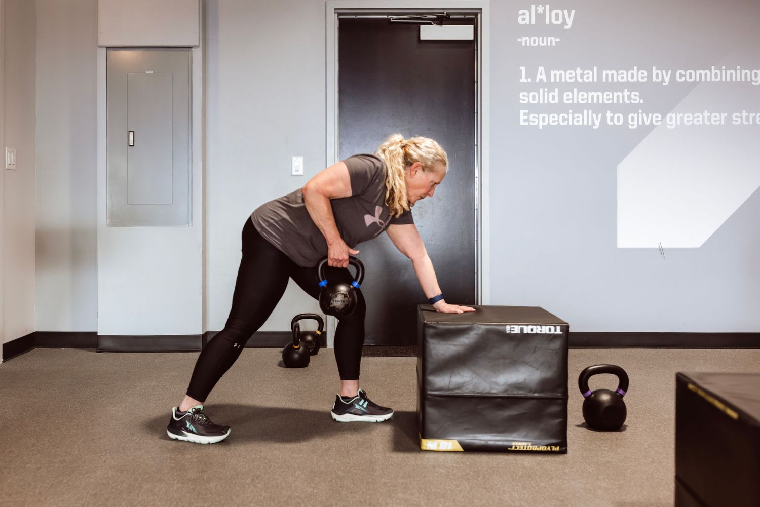 A person performs a low lunge exercise position on a gray exercise mat in a bright room.