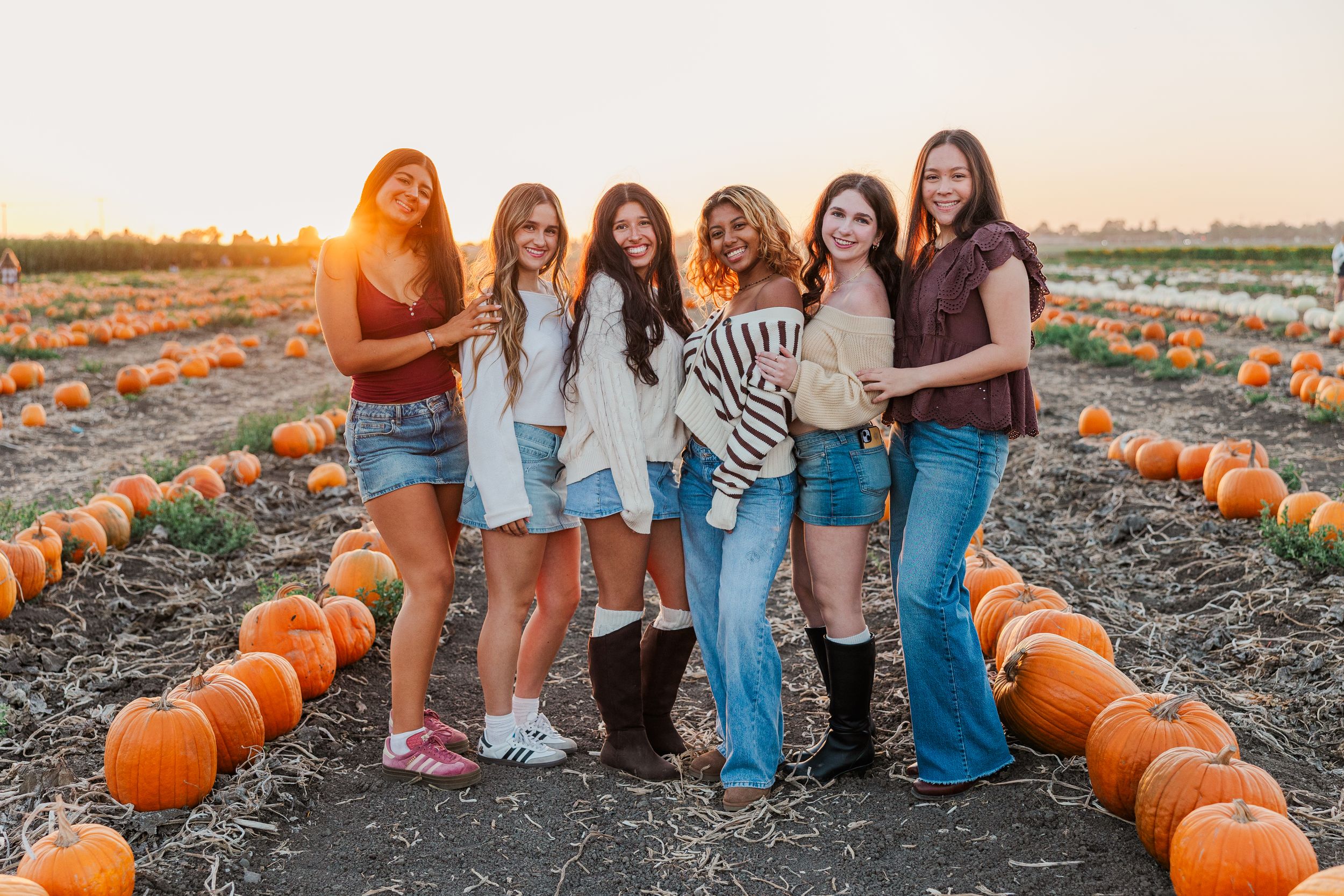 A group of friends poses together at a pumpkin patch during golden hour sunset.
