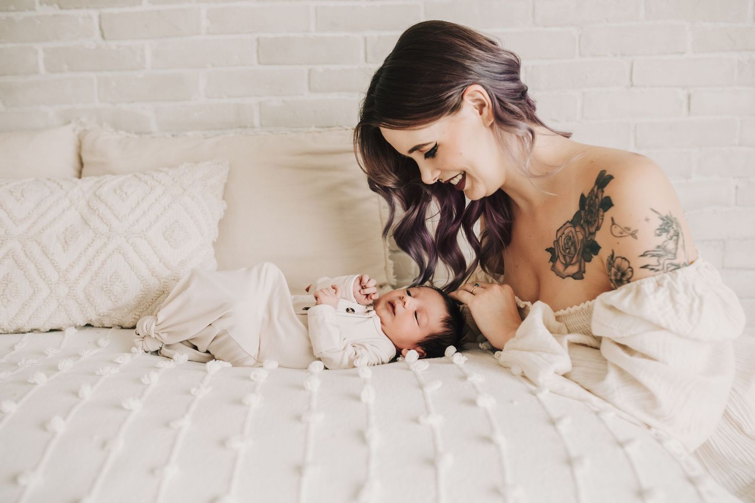 A mother laying over her newborn baby dressed in all white on white bedding.