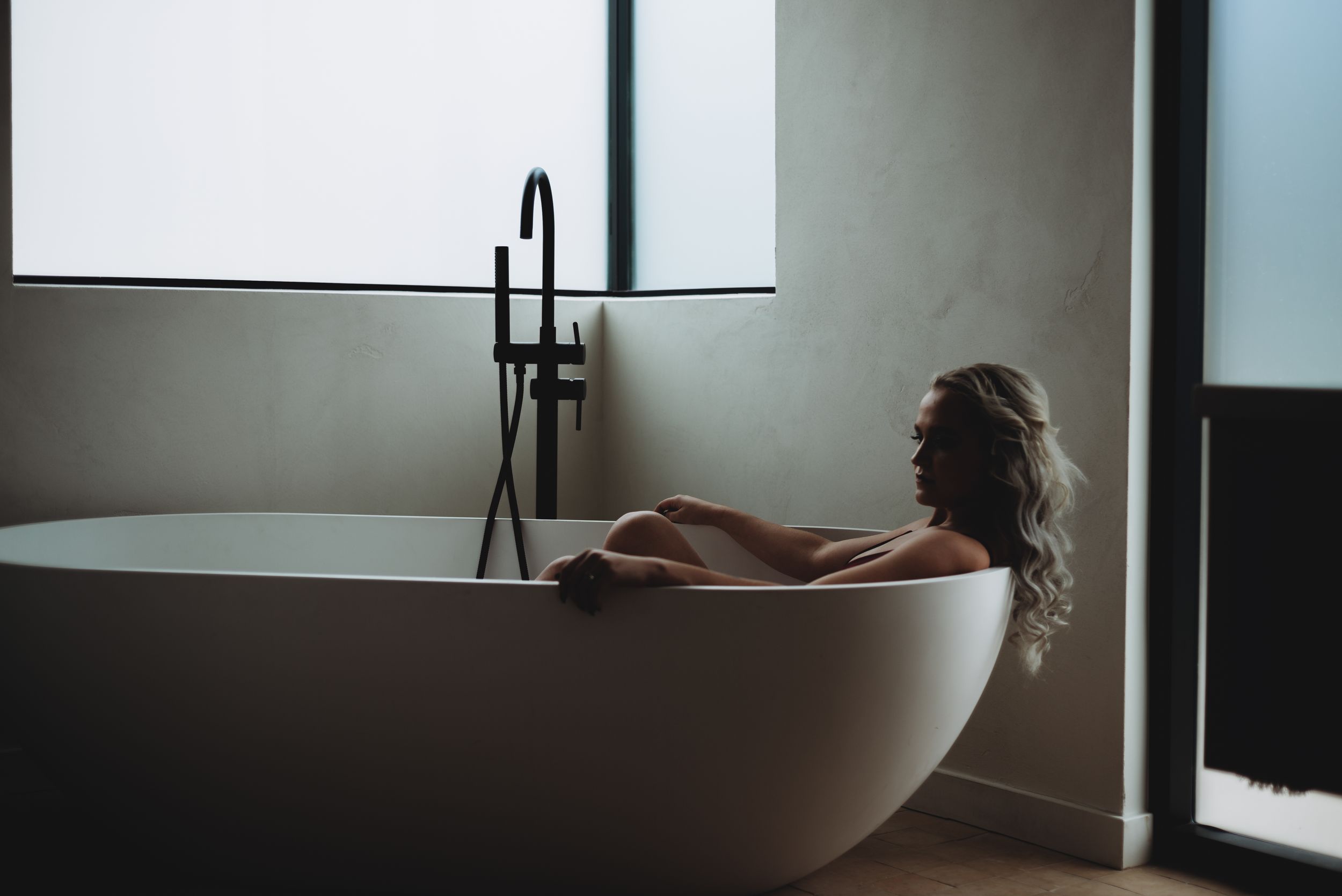 A sleek white freestanding bathtub sits in a modern bathroom with floor-to-ceiling windows and natural light.