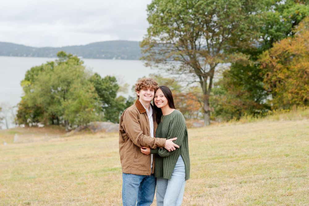 A couple embraces while standing in a golden autumn field with mountains and water visible in the distance.