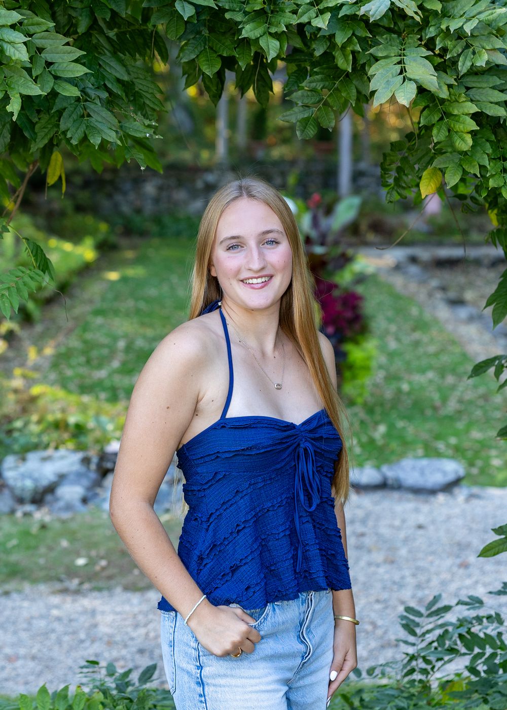 High school senior in blue top and jeans poses outdoors in an lush Italianate garden during yearbook portraits in Lincoln, MA