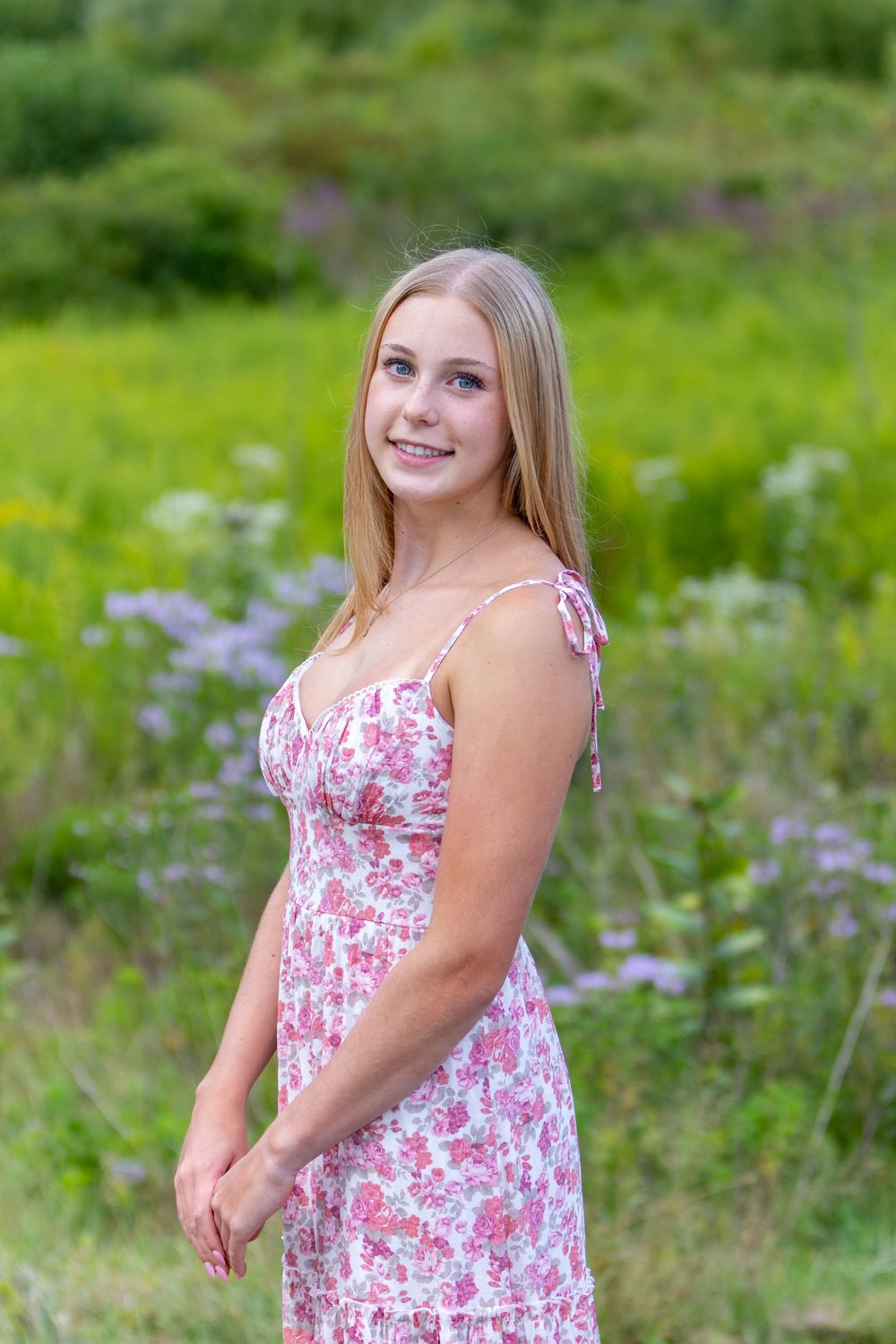 High school senior in a pink floral sundress poses outdoors in a field of purple wildflowers on a sunny summer day during yearbook portraits