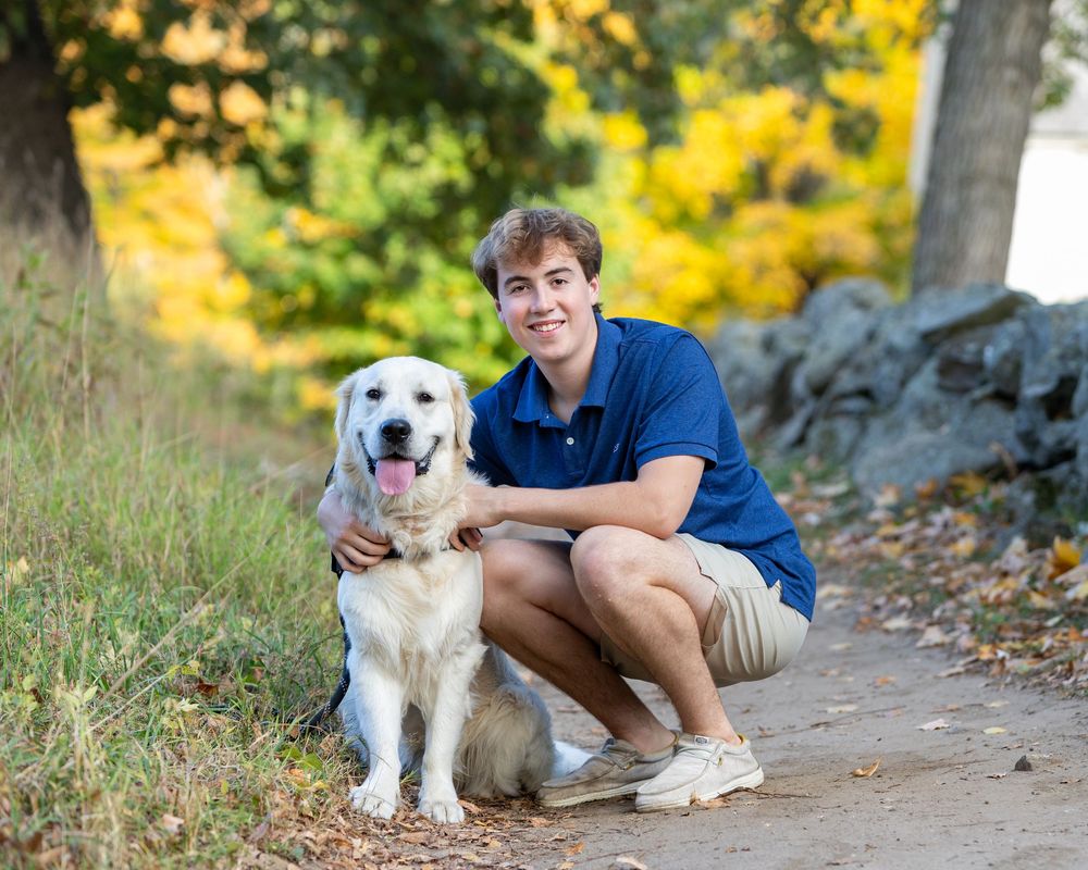 High school senior poses with a golden retriever dog with autumn foliage in background in Concord, MA for high school yearbook portraits.