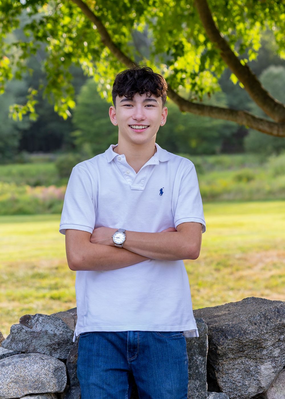High school senior in a white polo shirt and jeans sits casually on a stone wall with lush green trees in the background in Lincoln, MA.