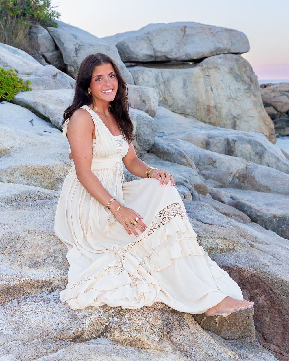 High school senior in a flowing white dress sits gracefully on coastal rocks during yearbook photoshoot Wingaersheek Beach- Gloucester, MA